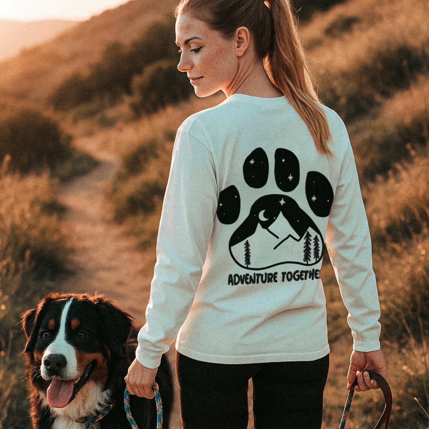 Frau wandert mit Bernersennenhund im Abendlicht. Sie trägt ein weißes Longsleeve aus nachhaltiger Bio-Baumwolle mit großem Pfotenabdruck und Naturmotiv auf dem Rücken. Der Schriftzug „ADVENTURE TOGETHER“ betont das gemeinsame Abenteuer.