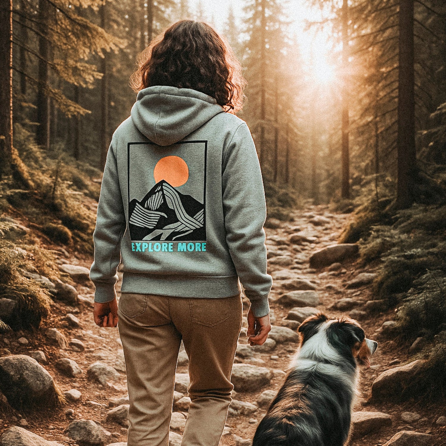 Frau wandert mit Australian Sheperd im Morgenlicht. Sie trägt einen grauen Hoodie aus nachhaltiger Bio-Baumwolle mit Bergprint und Naturmotiv auf dem Rücken. Der Schriftzug „Explore More“ betont das Abenteuer.